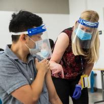 Clinard, wearing a face mask and face shield, points to a student&#039;s arm as another holds a needle.
