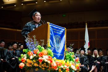 Komoto dressed in regalia at podium, speaking.
